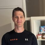 Mann in schwarzem Sportoberteil mit Logo eines Fitnessunternehmens, lächelnd vor einem Regal mit gesunden Lebensmitteln.