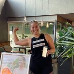 Frau präsentiert stolz ihre Muskeln und hält ein Handy mit Fitnessprogramm für Ernährungsfirma.