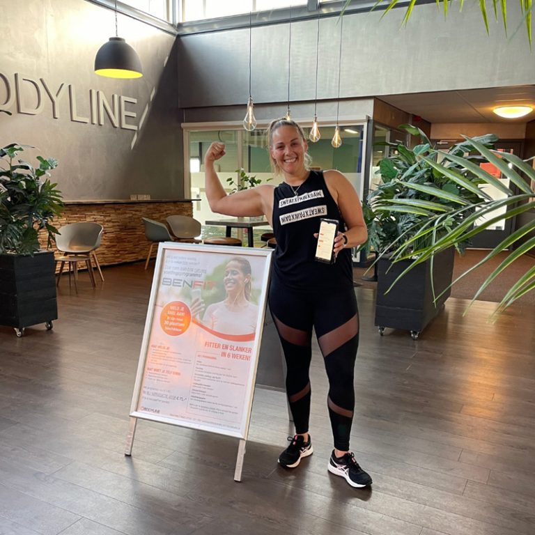Eine Frau präsentiert stolz ihre Muskeln vor einem Flyer, der für ein Fitnessprogramm wirbt.