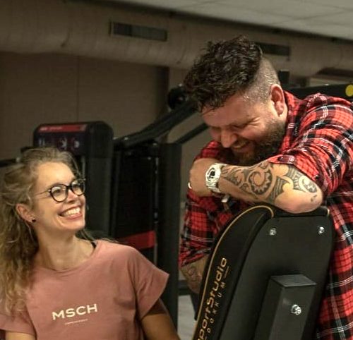 Fitnesspärchen lacht im Studio: Mann mit Tattoos und Frau mit Brille.