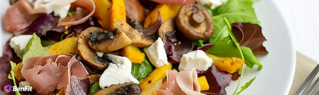 Nahaufnahme einer gesunden und sausgewogenen Mahlzeit, die zarte sScheiben Schinken, cremigen Ziegenkäse, geröstete Pilze und leuchtend orangefarbenen Kürbis über einem Bett aus frischen grünen und roten Blättern präsentiert.